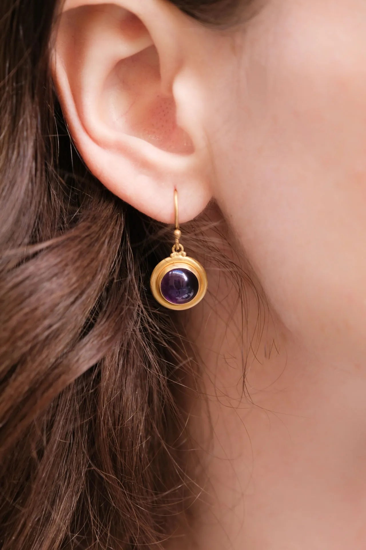 Gold earring with a purple gemstone worn by a person with brown hair.